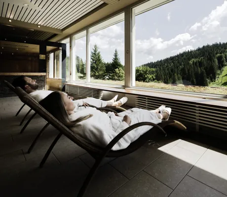 Paar in weißen Bademänteln liegt auf Ruheliegen vor dem Panoramafenster im Waldhotel am Notschreipass