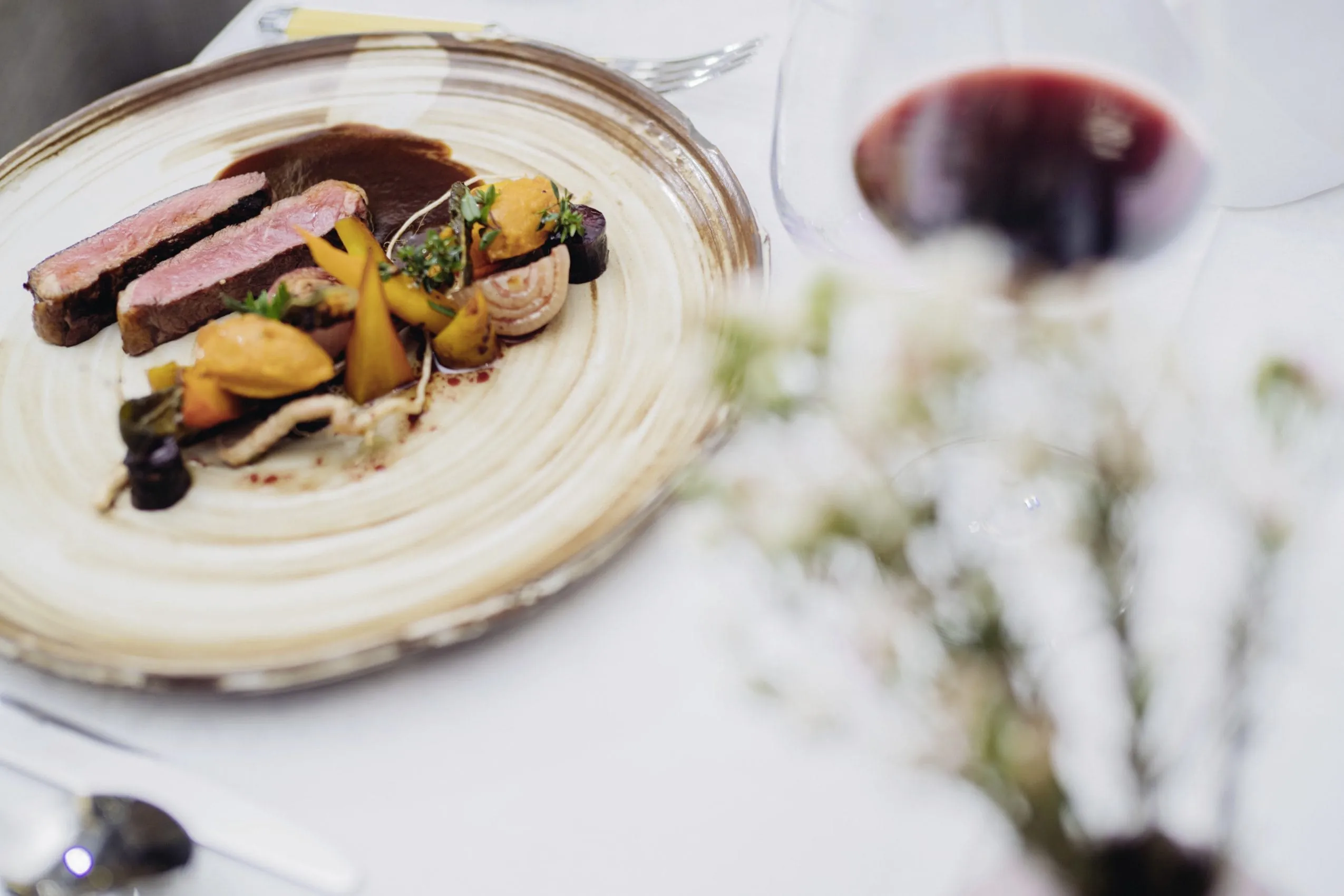 Fein angerichtetes Gericht des Reiters Supreme mit zwei rosa gebratenen Scheiben Fleisch, Gem&uuml;se, dunkler So&szlig;e und einem Glas Rotwein.