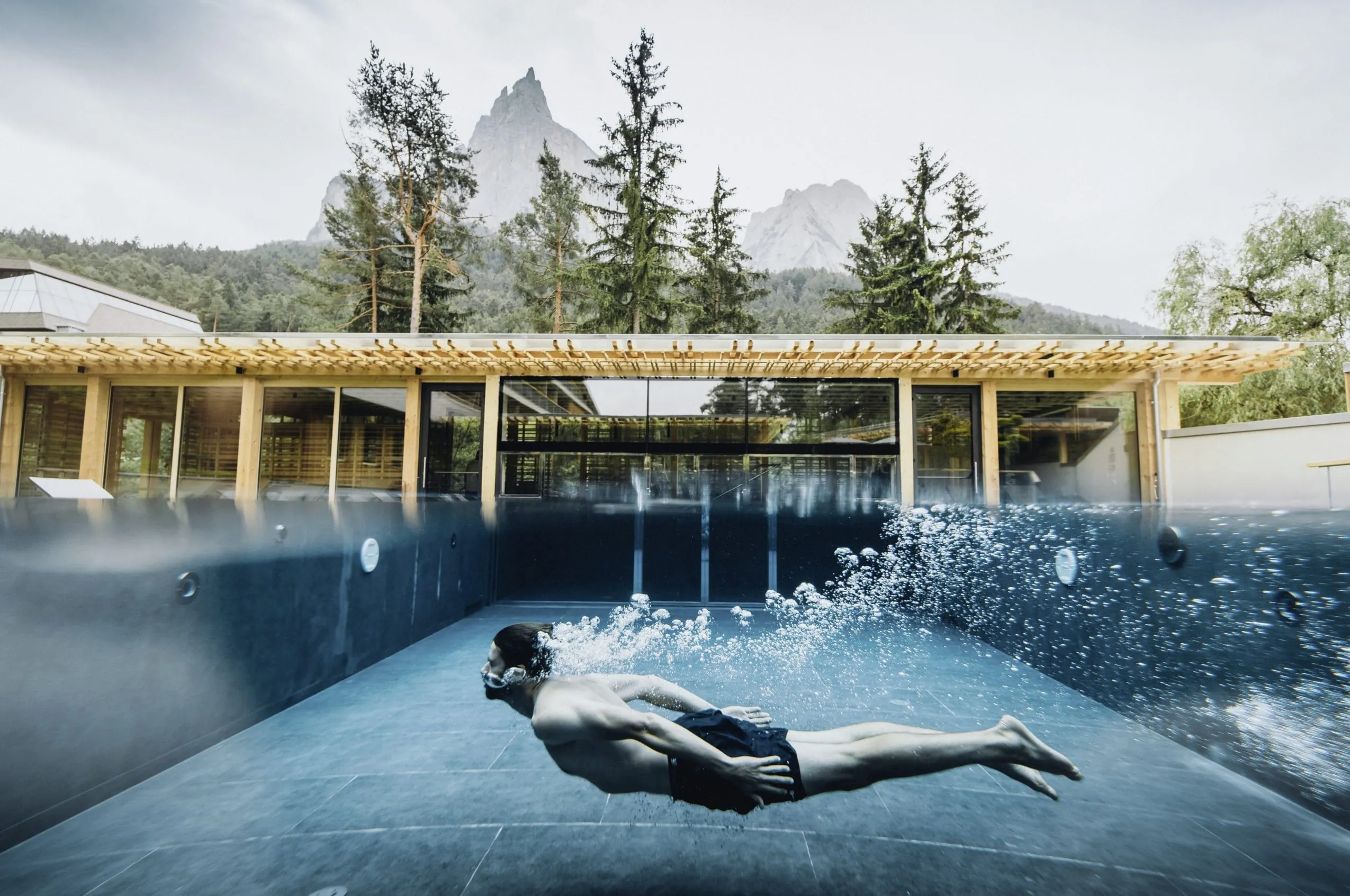 Junger Mann unter Wasser im Outdoorpool des Sensoria Dolomites. Im Hintergrund die Berge der Dolomiten