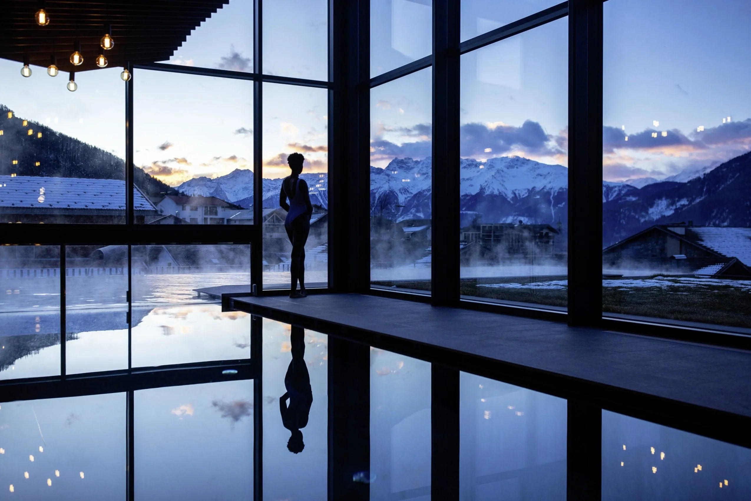 Frau steht an den gro&szlig;en Panoramafenstern des Indoorpools des Hotels Weisses Kreuz mit wundersch&ouml;nem Ausblick auf die S&uuml;dtiroler Berge bei D&auml;mmerung
