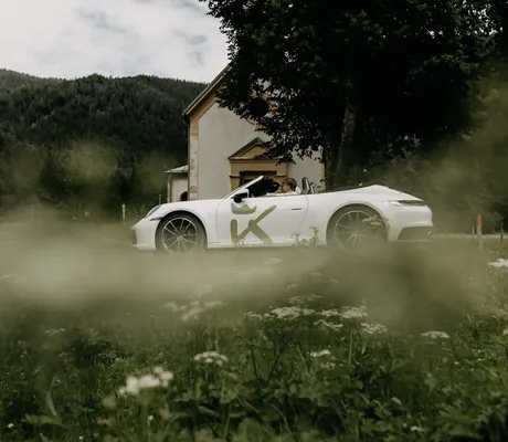 weißer Porsche mit Logo des Das Kronthaler hinter Wiese