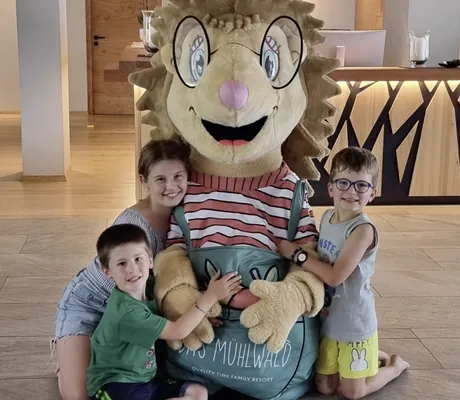 Drei Kinder umarmen freudig das Igel-Maskottchen im Familienhotel Das Mühlwald