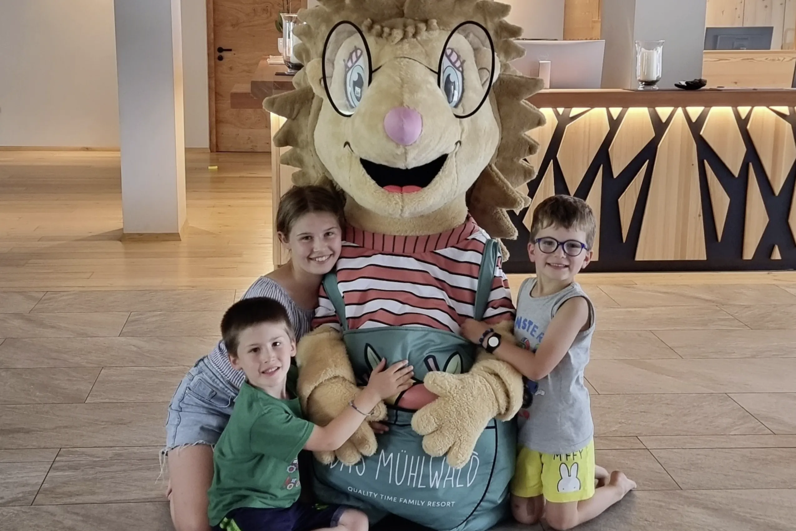 Drei Kinder umarmen freudig das Igel-Maskottchen im Familienhotel Das Mühlwald