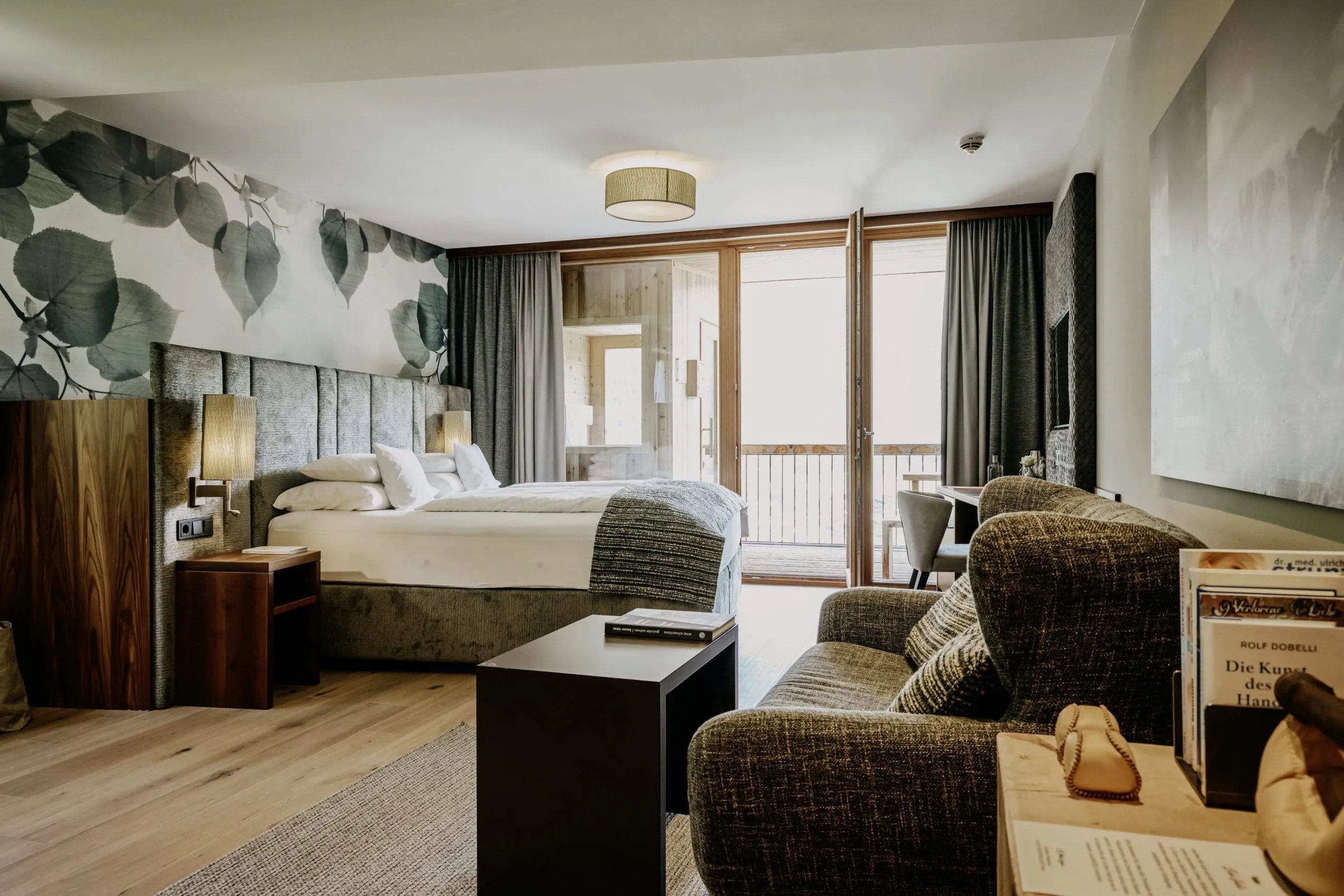 Zimmer des "DAS KRONTHALER" in Triol mit gro&szlig;em Bett mit grauem Polsterkopfteil, gro&szlig;en dunkelgr&uuml;nen Bl&auml;ttern an der Wand und Balkon.