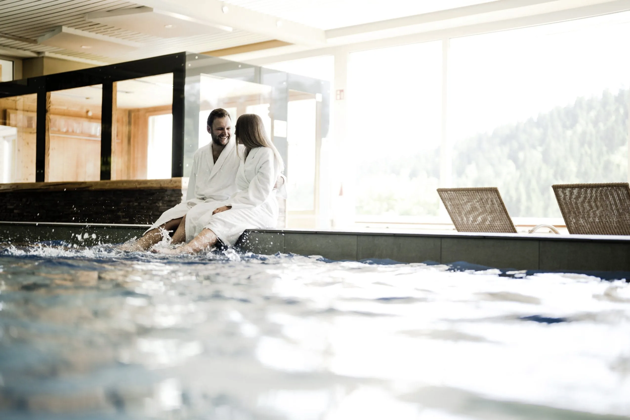 Am Beckenrand des Hallenbades im Waldhotel am Notschreipass sitzt ein Paar in wei&szlig;en Badem&auml;nteln, das sich anlacht.