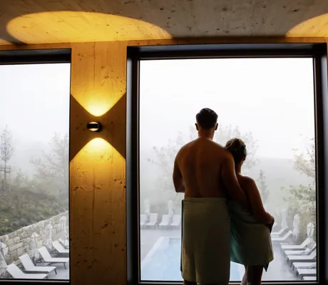 Pärchen steht im Spabereich und blickt aus den großen bodentiefen Fenstern nach draußen auf die Terrasse mit Outdoorpool.