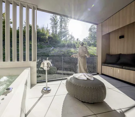 Frau in Bademantel steht am Balkonrand neben sprudelndem Whirlpool und schaut in die Landschaft