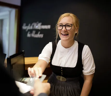 blonde Frau im Dirndl an der Rezeption übergibt Zimmerkarte an Gast in Haubers Naturresort