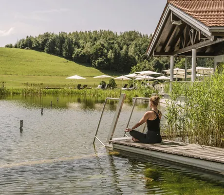 Frau in schwarzem Fitnessoutfit macht Yoga auf Holzsteg des Natursees in Haubers Naturresort