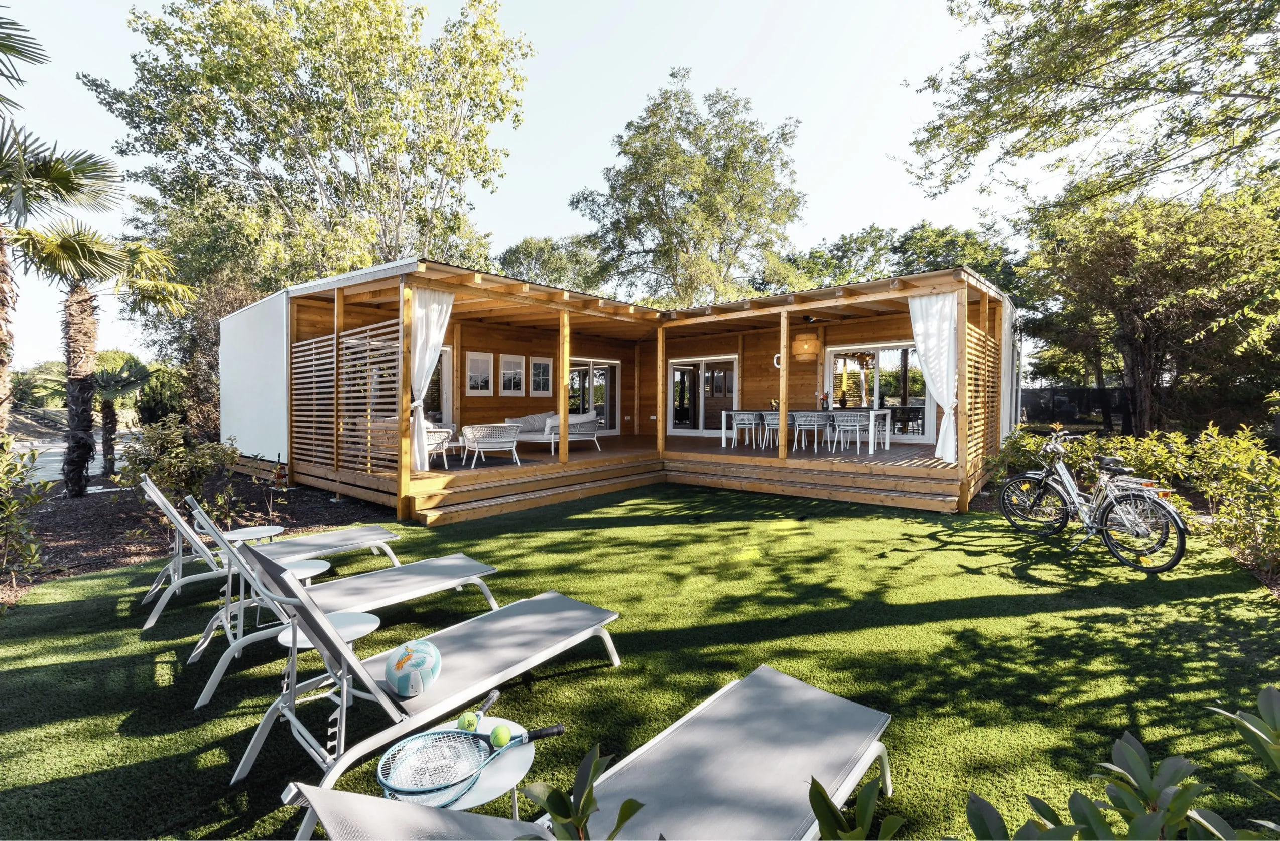Blick auf die Cottage Villa aus Holz des Tenuta Primero Grado Family Collection mit möblierter Terrasse und großem Garten mit vier Sonnenliegen und Fahrrädern