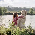 Junges Mädchen legt ihren Arm auf Golden Retriever, beide blicken auf einen idyllischen See, umgeben von grüner Natur