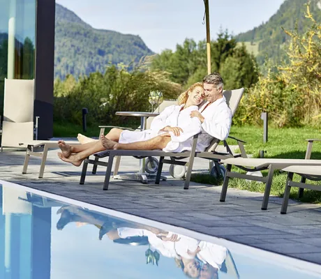 Paar in weißen Bademänteln liegt gemeinsam auf einer Sonnenliege am Outdoorpool des Hotel Oberstdorf