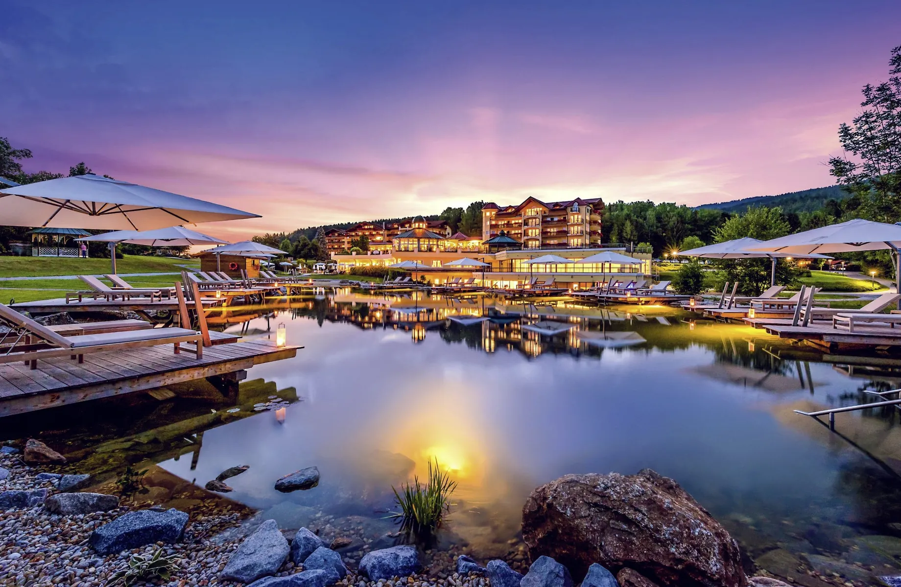 Beleuchtete Außenansicht des Hotel Mooshof Wellness & Spa Resort am Abend mit pink gefärbtem Himmel und Naturbadeteich im Vordergrund
