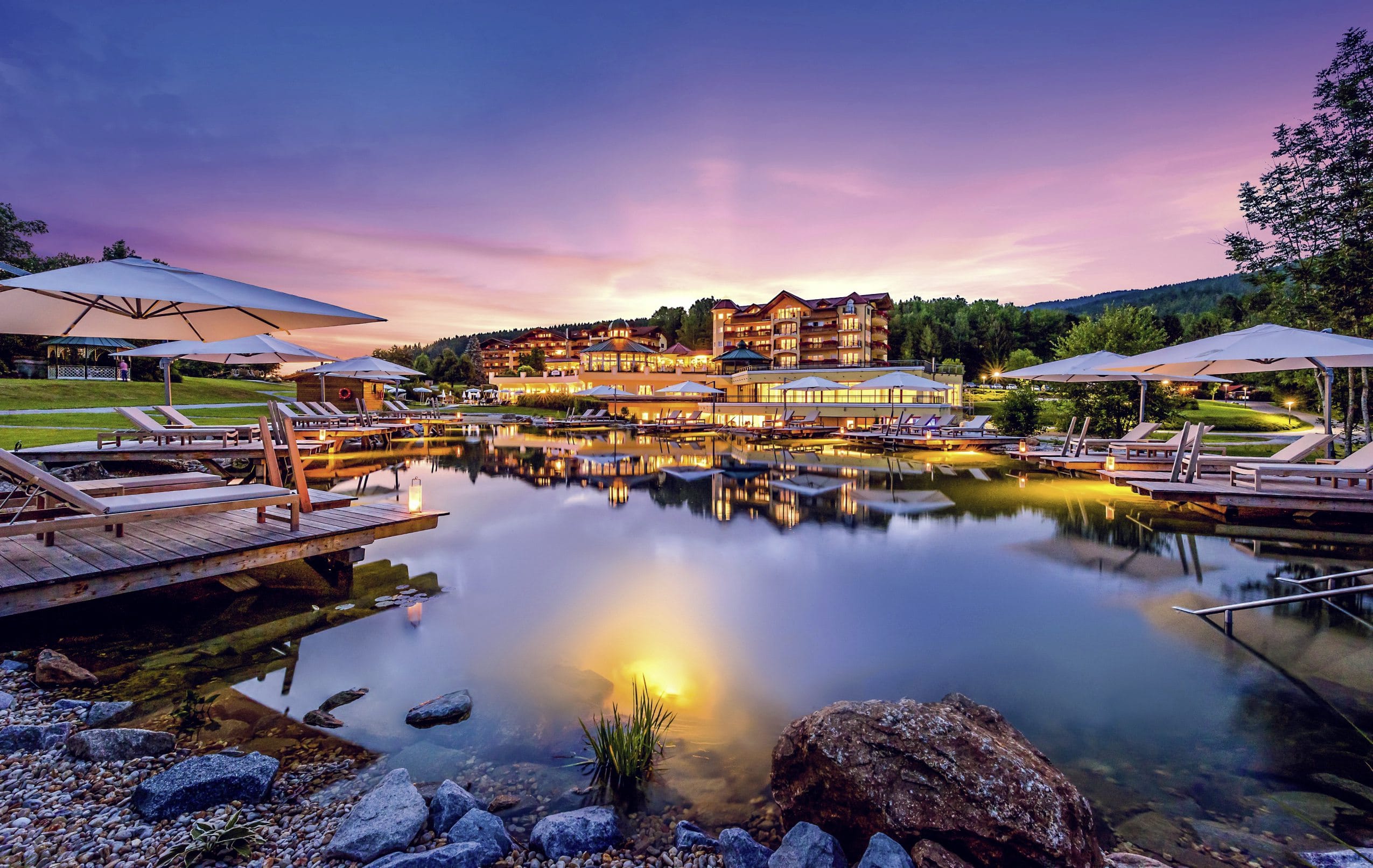 Beleuchtete Außenansicht des Hotel Mooshof Wellness & Spa Resort am Abend mit pink gefärbtem Himmel und Naturbadeteich im Vordergrund