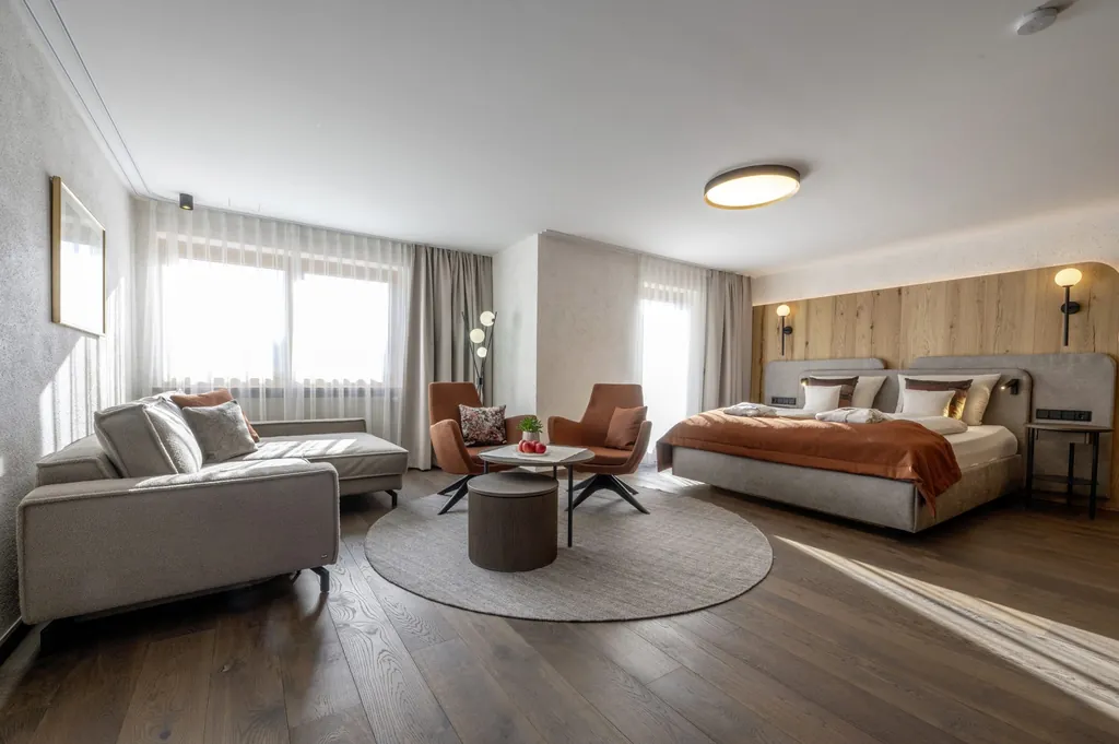 Blick in das Doppelzimmer des Hotel Mooshof Wellness & Spa Resort mit großem Doppelbett, orange-rötlichen Sesseln und einem hellgrauen Sofa