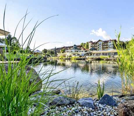 Blick auf den idyllischen Naturbadeteich des Hotel Mooshof Wellness & Spa Resort mit mehreren Sonnenliegen und -schirmen ringsherum