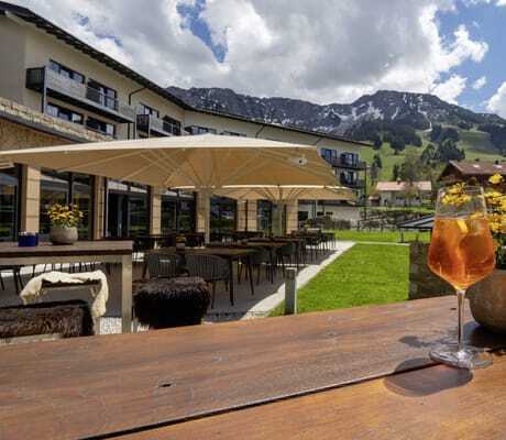 Ein Glas Aperol auf dem Terrassentisch des Panoramahotels Oberjoch im Frühling mit Blick auf die Hotelfassade und die Berge