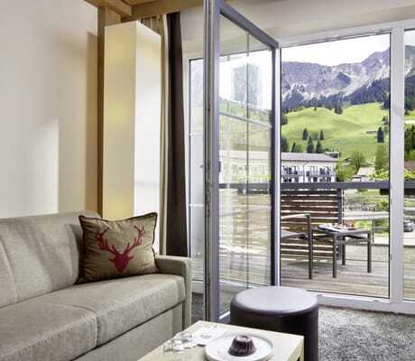 hellgraues Sofa mit Hirschgeweih-Kissen im Zimmer des Panoramahotel Oberjoch mit Balkon und Blick auf die grünen Berge im Sommer