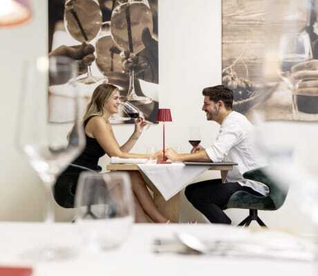 Paar sitzt am Tisch vor Fotos aus Südtirol im Restaurant des Das Parkhotel und trinkt lachend Rotwein