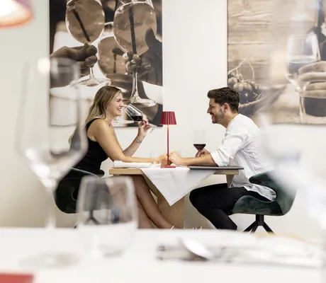 Paar sitzt am Tisch vor Fotos aus Südtirol im Restaurant des Das Parkhotel und trinkt lachend Rotwein