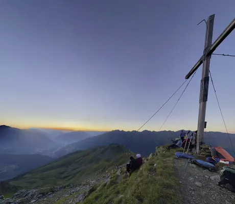 Blick vom Gipfel mit großem Holzkreuz, steinigem Pfad, Grasflächen und mehreren Personen beim Sonnenaufgang, Panorama aus Tälern und Bergketten im Hintergrund in der Nähe des Das SeeMOUNT – Active Nature Resort ****Superior