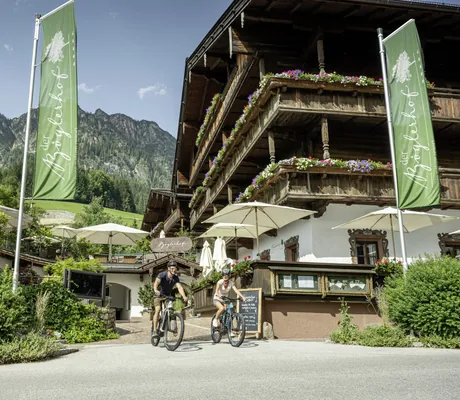 zwei Fahrradfahrer vor der Fassade des Der Böglerhof – pure nature spa resort neben zwei grünen Fahnen