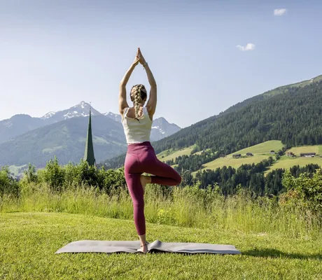 Eine Frau macht Yoga-Übung im Garten des Der Böglerhof – pure nature spa resort mit Blick auf Berglandschaft und Kirchturm