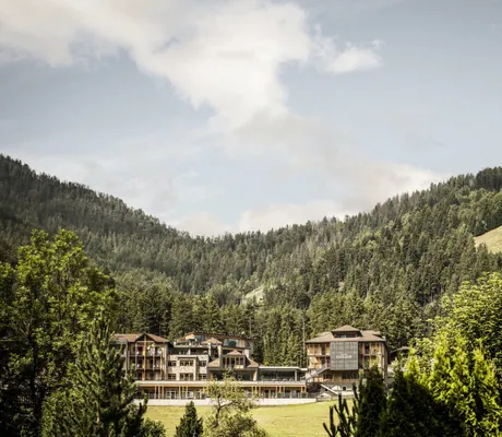 Blick auf die Fassade des Excelsior Dolomites Life Resort umgeben von grünen Bergen und Wäldern