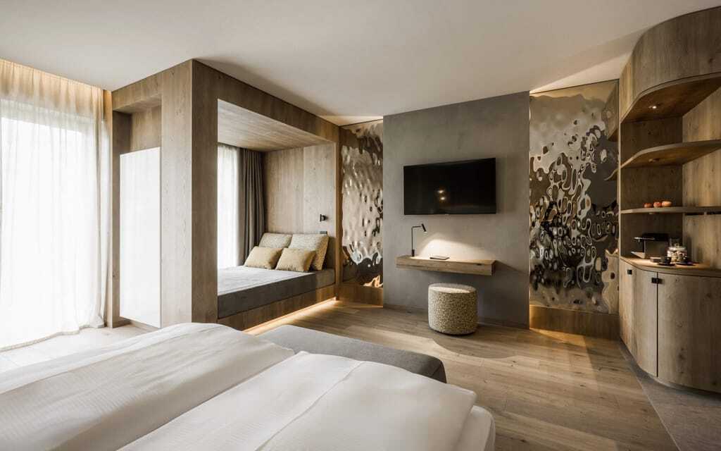 Zimmer im Excelsior Dolomites Life Resort mit viel Holzelementen und neutralen Farben, Doppelbett, Flachbild-Fernseher, gemütlicher Sitzbank und kleiner Küchenzeile mit Kaffeemaschine