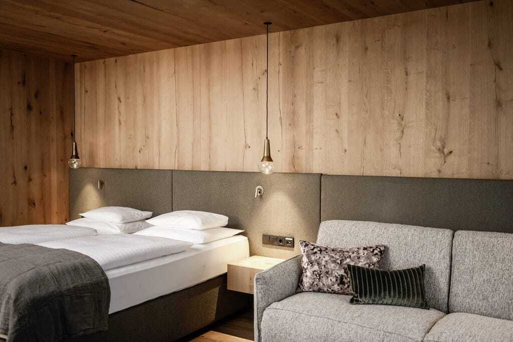 Modernes Schlafzimmer mit natürlicher Holzvertäfelung, stilvoller Beleuchtung, gemütlichem Doppelbett und hellgrauem Sofa im Peternhof Wohlfühlresort