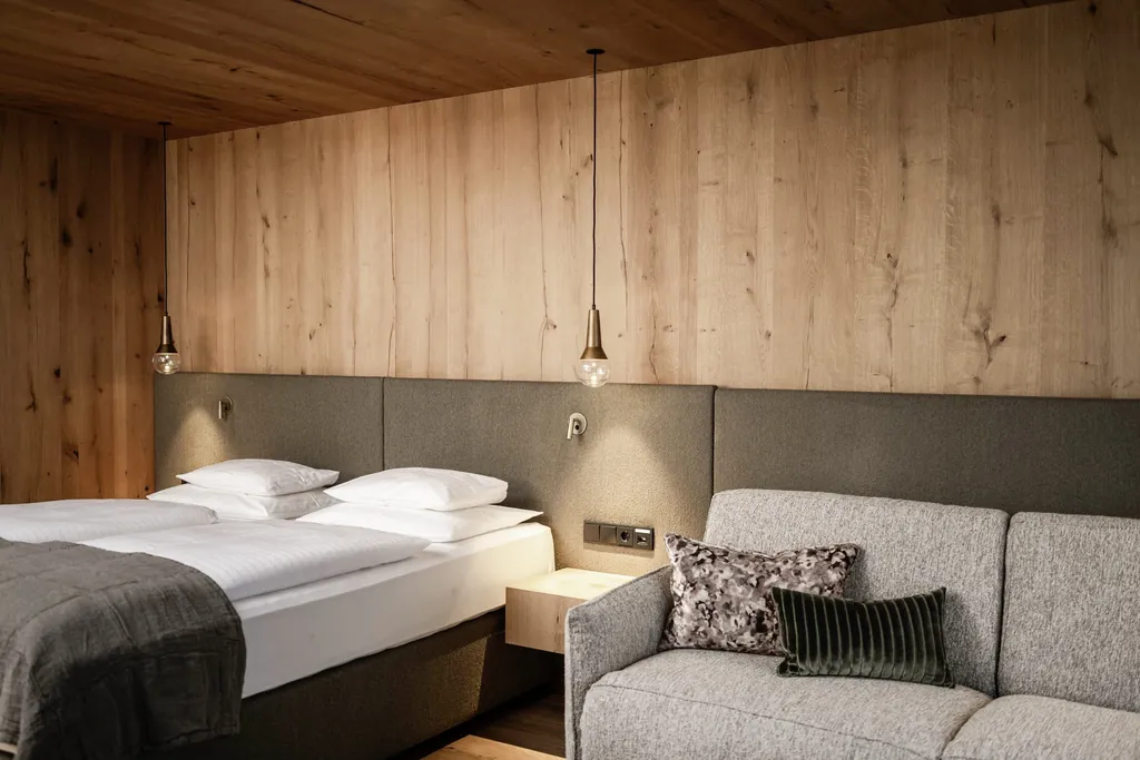 Modernes Schlafzimmer mit natürlicher Holzvertäfelung, stilvoller Beleuchtung, gemütlichem Doppelbett und hellgrauem Sofa im Peternhof Wohlfühlresort
