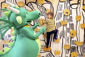 Drachen-Maskottchen des Familotel Elldus Resort steht neben einem Jungen, der eine Kletterwand hinaufklettert
