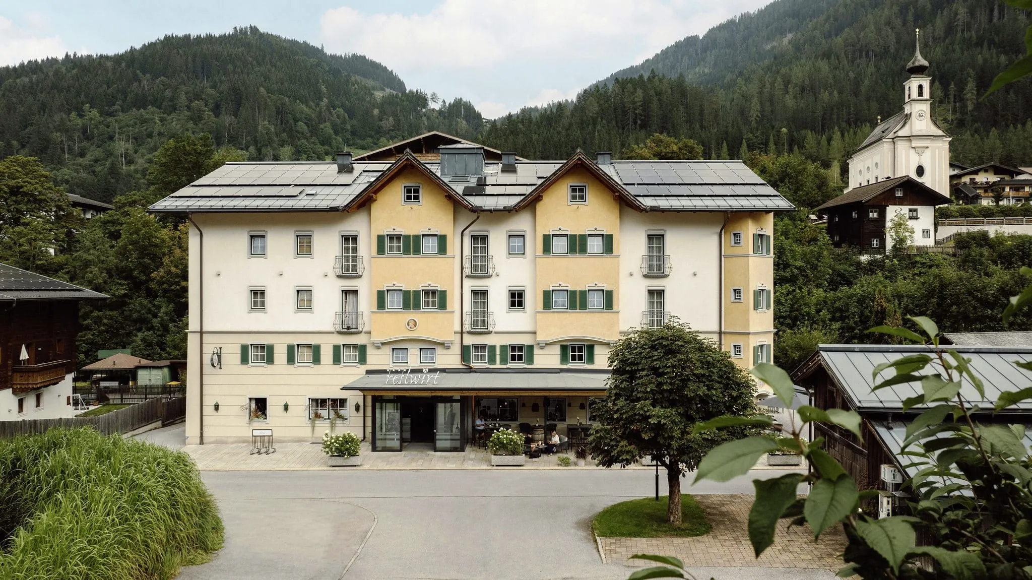 Fassade des Resl Resort im traditionellen Stil, eingebettet in eine malerische Berglandschaft und Kirche im Hintergrund