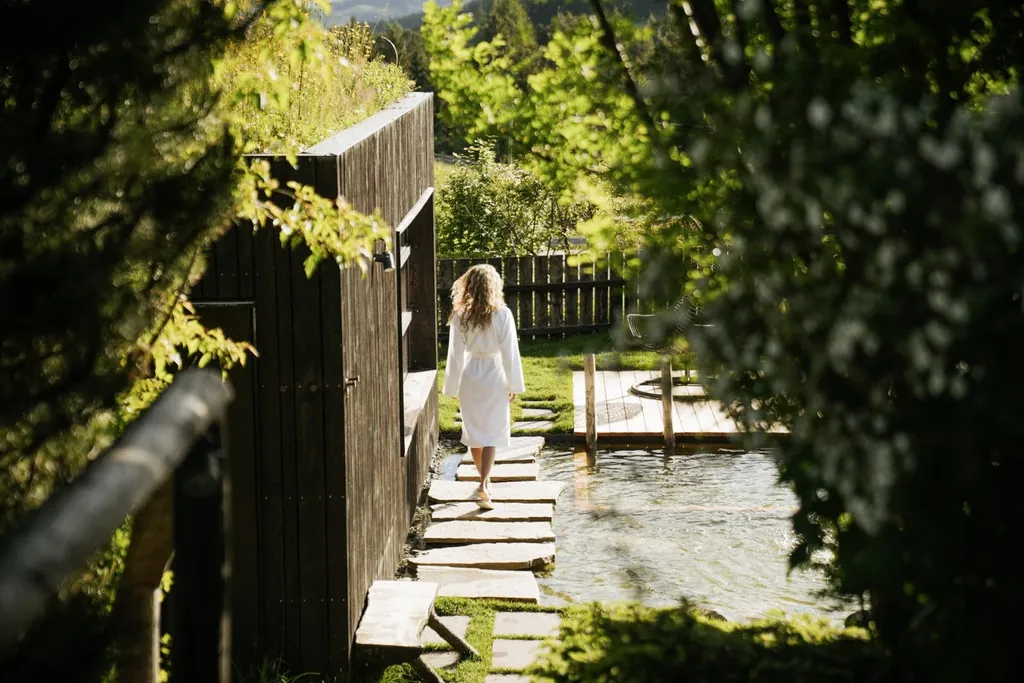 Frau im Bademantel läuft über große Steinplatten durch den üppig grünen Wellnessgarten mit Naturbadeteich im Naturhotel Pfösl