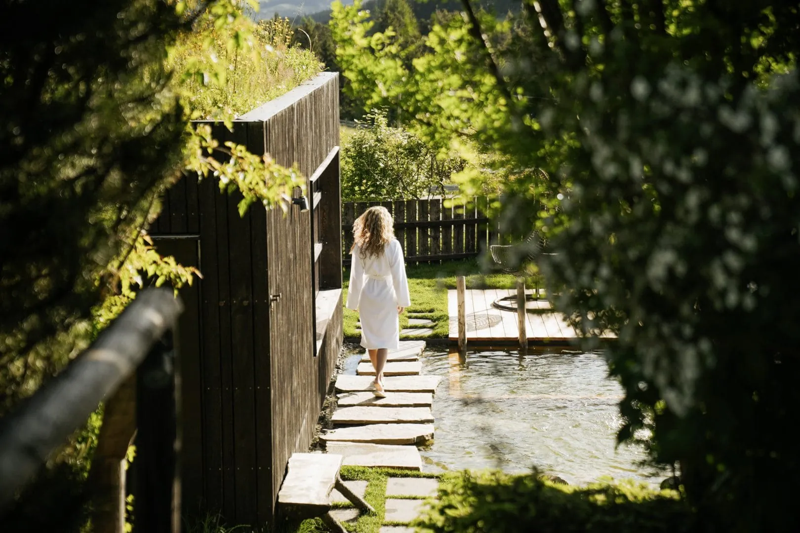 Frau im Bademantel läuft über große Steinplatten durch den üppig grünen Wellnessgarten mit Naturbadeteich im Naturhotel Pfösl