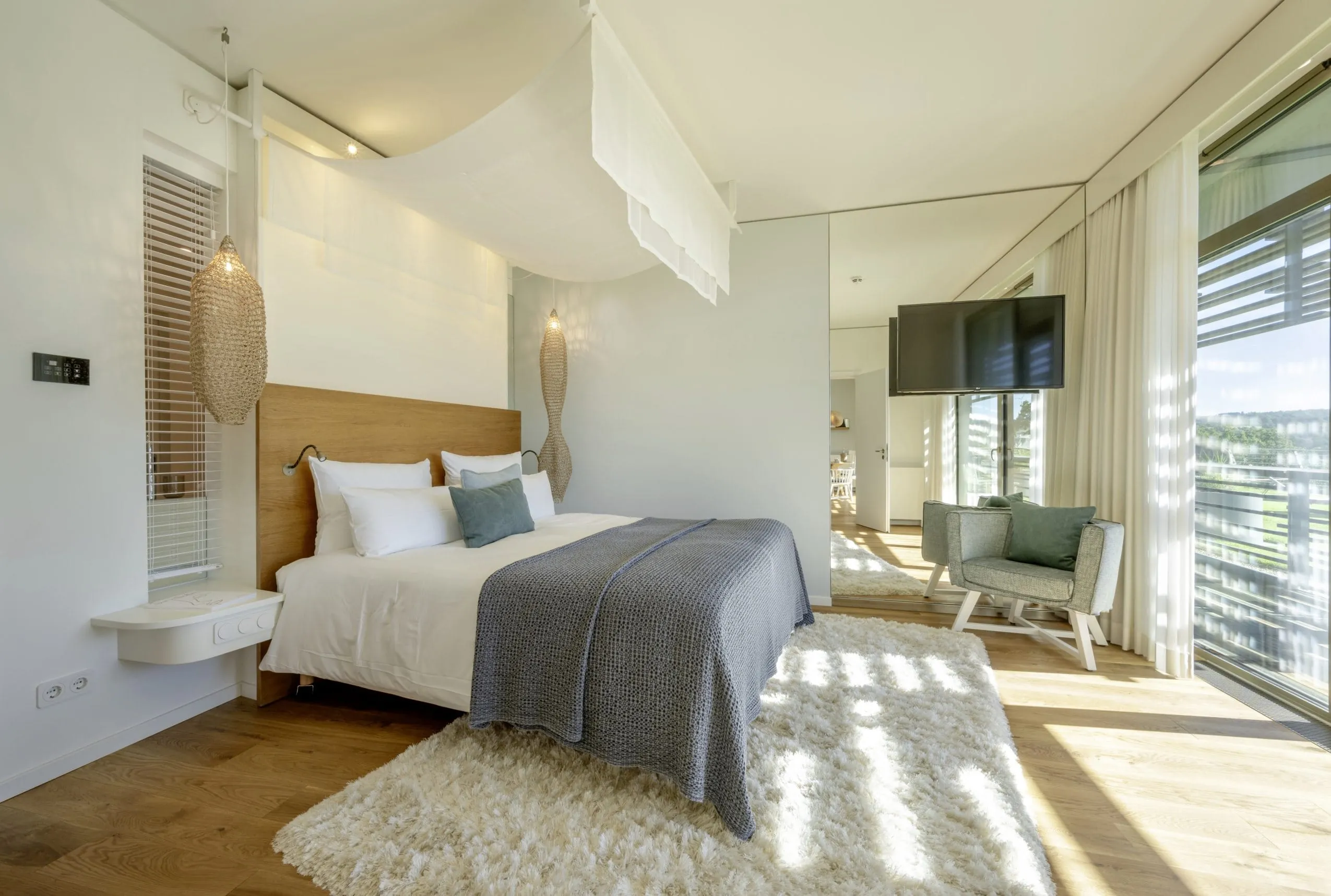 Helles Hotelzimmer mit Holzbett, grauen Textilien, Designer-H&auml;ngeleuchten und bodentiefen Fenstern mit Naturblick