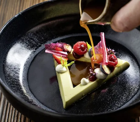 Hand gießt Schokosauce auf ein Dessert des Der Böglerhof – pure nature spa resort aus Waldmeistercreme, verschiedenem Obst und Sauerrahm