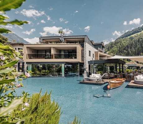 Moderne, luxuriöse Anlage der Quellenhof See Lodge mit Naturholz-, Glas- und Steinelementen, verschiedenen Pools, hölzerne Plattformen im Wasser mit Liegen, ein Boot und eine malerische Bergkulisse im Hintergrund