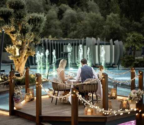 Paar beim Dinner auf einem beleuchteten Holzdeck über Wasser in der Quellenhof See Lodge, dekoriert mit Lichterketten, Kerzen und Blumen, beleuchtete Wasserfontänen im Hintergrund