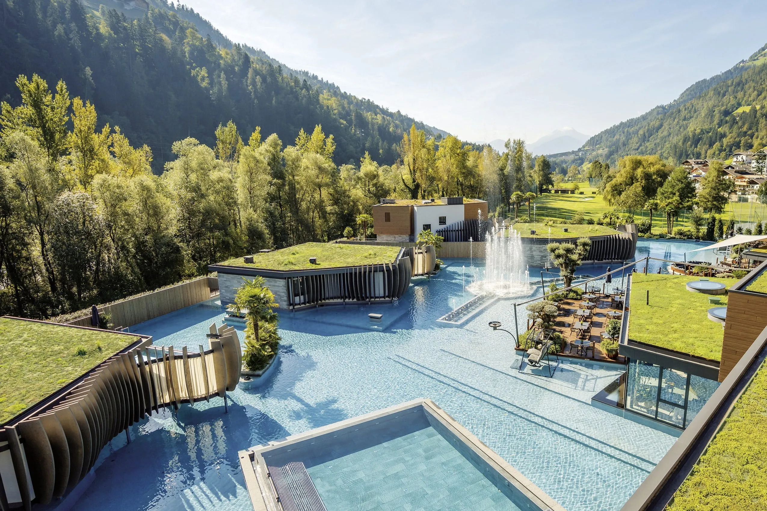 Blick auf die große Poolanlage der Quellenhof See Lodge mit Springbrunnen, grünen Dächern und möblierter Holzterrasse