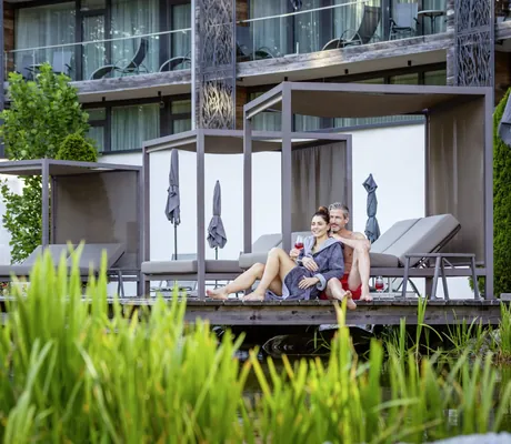 Paar entspannt sich auf einer Holzterrasse im Außenbereich des Wellness & Naturresort Reischlhof mit gemütlichen Daybeds im Hintergrund