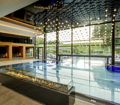 Innenpool des Wellness- und Naturresort Reischlhof mit großer Fensterfront, moderner Beleuchtung, Liegen und Blick auf grüne Außenanlage