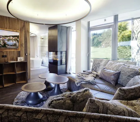 Gemütliches Zimmer im Wellness- und Naturresort Reischlhof mit großer grauer Eckcouch, drei runden Couchtischen und modernem Kamin in der Mitte, große Fenster mit Blick auf grüne Landschaft