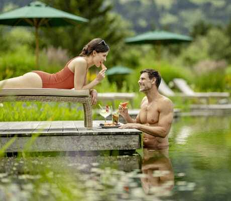 Frau in rotem Badeanzug auf einer Liege, Mann im Naturbadeteich des Sonnenalp Resorts, beide haben ein Stück Wassermelone in der Hand
