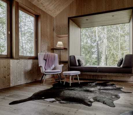 gemütlicher Wohnbereich im Baumhaus des Hofgut Hafnerleiten mit altrosafarbenem Sessel, Hocker, Stehlampe und braun, gepolsterten Sitzbank direkt am Panoramafenster mit Waldblick