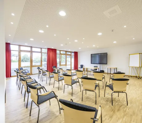 Stuhlreihen im Tagungsraum des Hotel Alpenblick Ohlstadt mit großem Monitor und orangen Vorhängen
