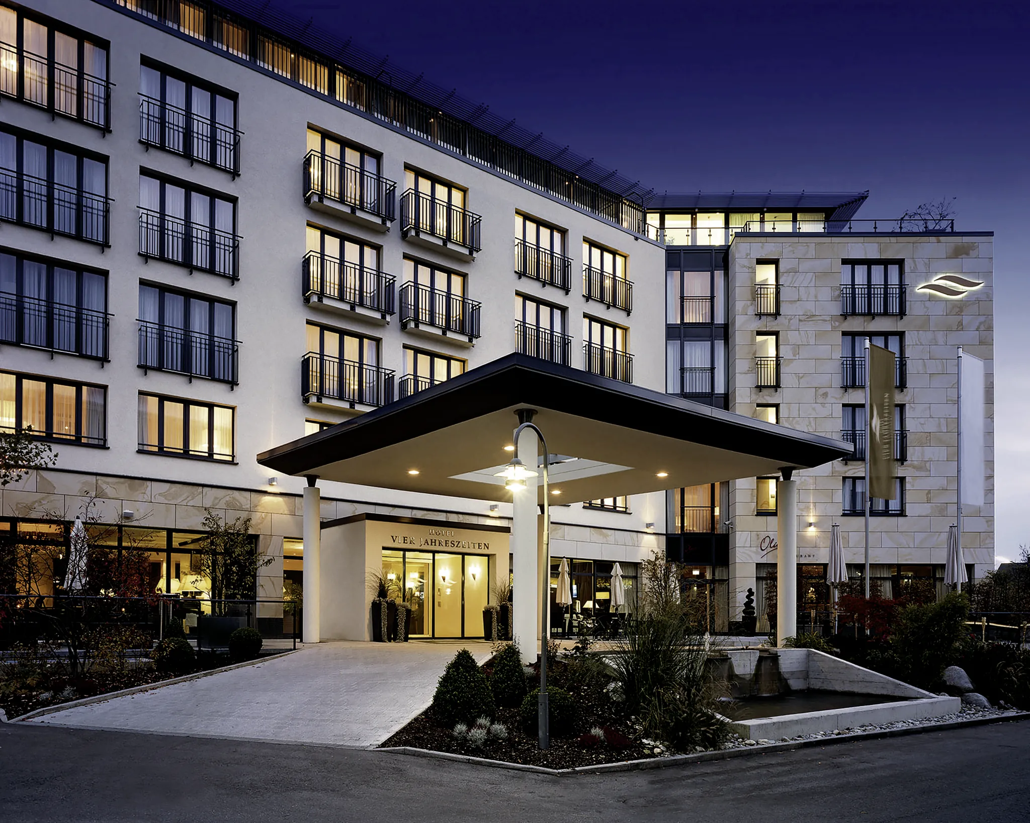Beleuchtete Hotelfassade mit Balkonen und Eingangsbereich des Hotel Vier Jahreszeiten Starnberg bei Abenddämmerung