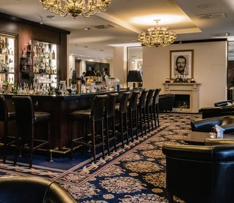 Elegante Hotelbar des Hotel Vier Jahreszeiten Starnberg mit dunklem Holz, Ledersesseln, Kronleuchtern und einem Kamin mit einem Porträt darüber