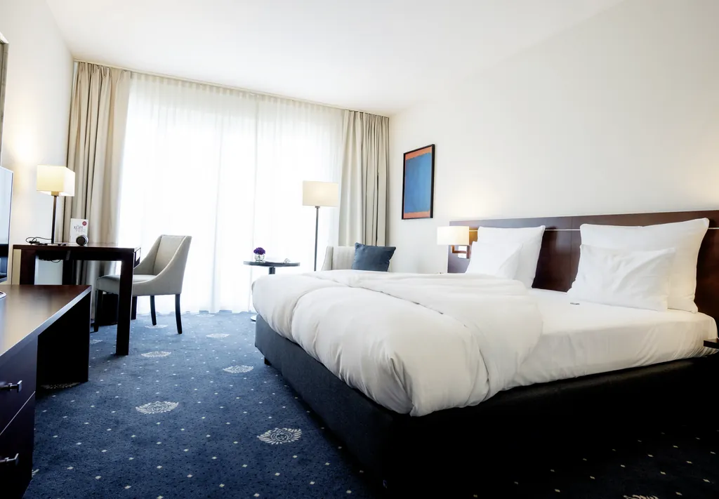 Helles Hotelzimmer des Hotel Vier Jahreszeiten Starnberg mit großem Doppelbett, Schreibtisch, Stuhl und bodentiefen Fenstern mit Vorhängen