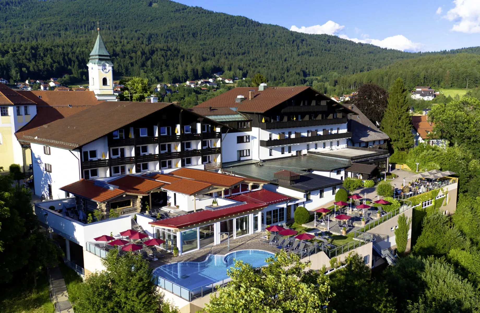 Außenansicht des Wellnesshotel Hofbräuhaus mit roten Sonnenschirmen und Außenpool, umgeben von grünen Bäumen und Bergen im Hintergrund